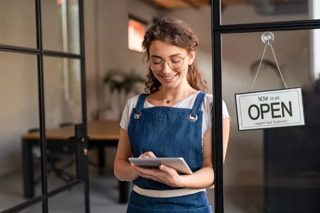 A woman in glasses, wearing a blue apron, smiling as she looks at a tablet, positioned near a door with an "Open" business sign, capturing a moment of business readiness, technology, and customer engagement.