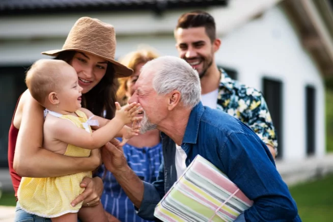 A heartwarming scene of a multigenerational family outside, with a baby's hands outreached towards an older man who's smiling, encapsulating a moment of connection, love, and the cycle of life in a natural environment.